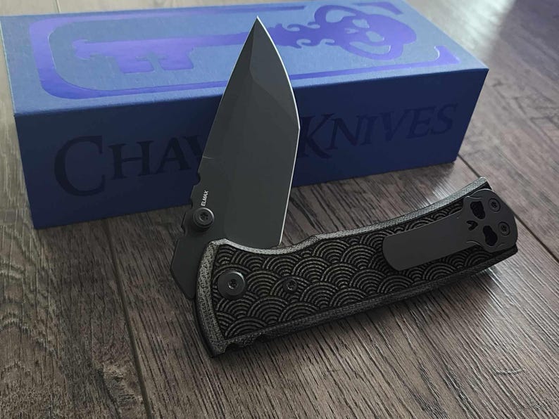 May include: A black folding knife with a partially open blade, resting on a wooden surface. The handle features a textured pattern and a pocket clip. A blue box with the text "CHAVES KNIVES" is in the background.