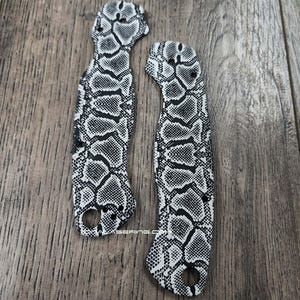 May include: Two knife handle scales with a black and white snakeskin pattern. The scales are made of a durable material and have a hole for a lanyard. The scales are designed to replace the original handles on a folding knife.