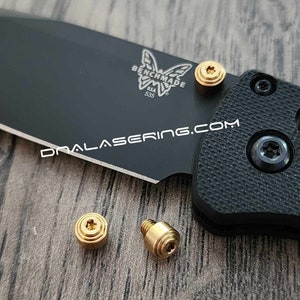 May include: Three gold-colored screws with a star-shaped head. The screws are on a wooden surface next to a black folding knife with the Benchmade logo and the text "USA 535".