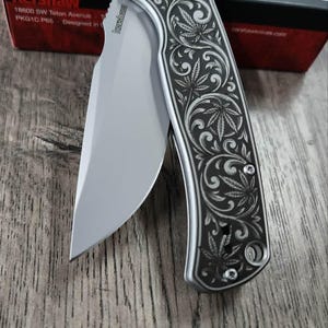 May include: A silver folding knife with a curved blade and a decorative handle featuring a leaf design. The knife is partially open, revealing the sharp edge. A red and black box is in the background.