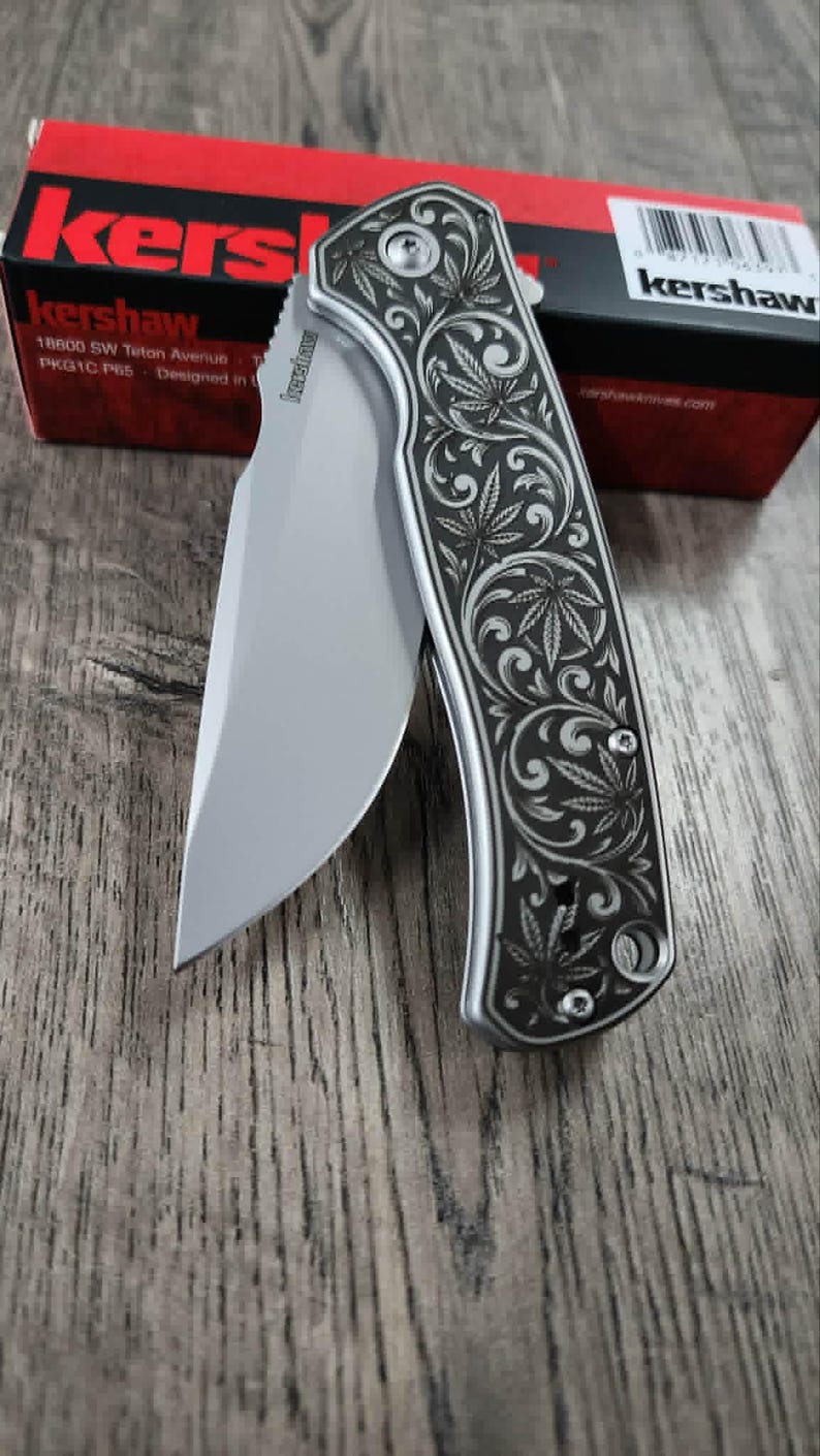 May include: A silver folding knife with a curved blade and a decorative handle featuring a leaf design. The knife is partially open, revealing the sharp edge. A red and black box is in the background.