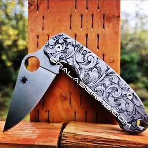 May include: A silver pocket knife with a decorative engraved pattern on the handle. The blade is closed and the knife is resting on a wooden surface.