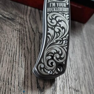May include: A silver and black pocket knife with detailed floral engraving. The handle displays a portrait and the text "I'M YOUR HUCKLEBERRY". The knife is partially open, showing the blade. Made in the USA.