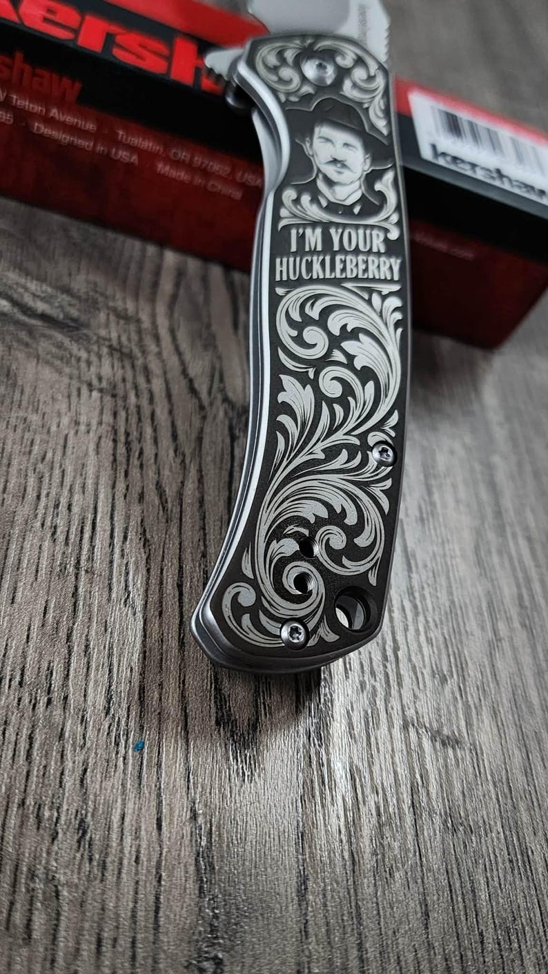 May include: A silver and black pocket knife with detailed floral engraving. The handle displays a portrait and the text "I'M YOUR HUCKLEBERRY". The knife is partially open, showing the blade. Made in the USA.