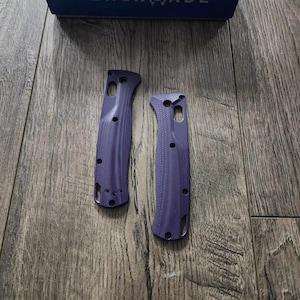 May include: Two purple knife handle scales with a textured surface, lying on a wooden surface. The scales are from a Benchmade Bugout knife. A blue box with the Benchmade logo is in the background.