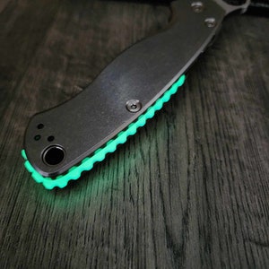 Peut inclure: Un couteau de poche noir avec une poignée verte qui brille dans le noir. Le couteau porte le logo Spyderco sur la poignée.