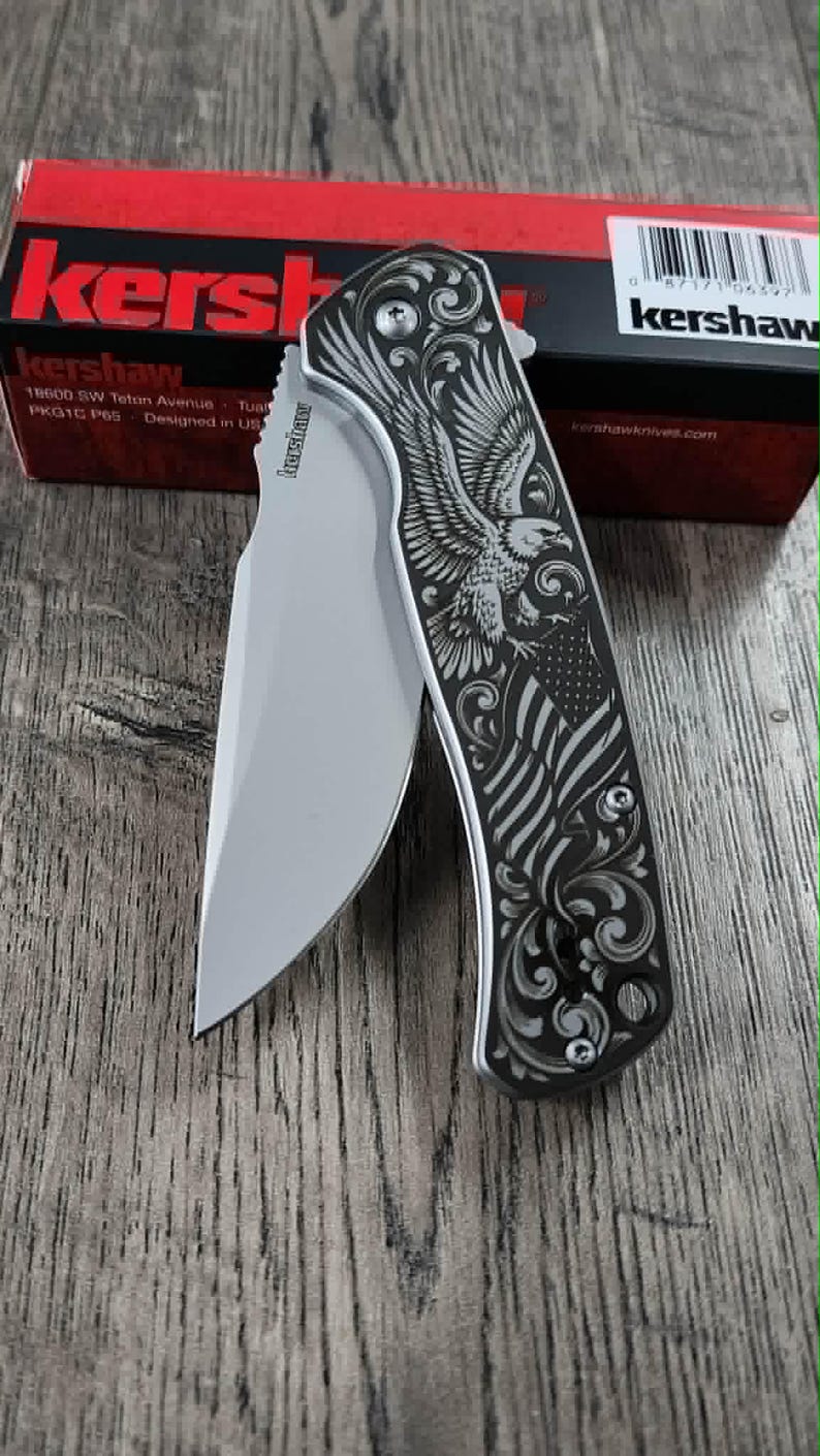 May include: A silver folding knife with a detailed engraving of an eagle and American flag on the handle. The knife blade is open, and the handle has a decorative floral design. The box in the background has the brand name "Kershaw".