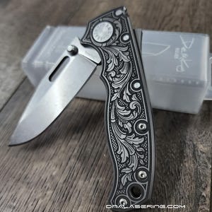 May include: A silver folding knife with an ornate floral pattern engraved on the handle. The blade is partially open, revealing a sharp edge. The knife is resting on a wooden surface, with a clear plastic case in the background.