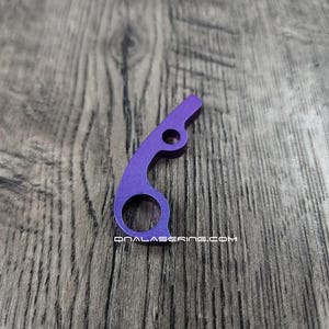 May include: A purple metal tool with a curved design, featuring a large circular opening and a smaller hole. The text "DNALASERING.COM" is printed on the tool. The tool is set against a wooden background.