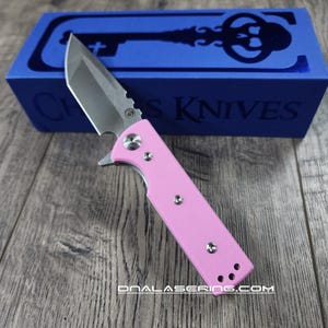 Può includere: Un coltellino tascabile rosa con una lama argentata e un logo a forma di chiave sulla scatola. La scatola è blu e dice "Chris Knives".