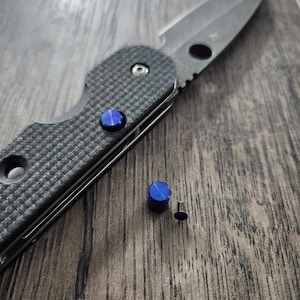 May include: A black folding knife with a blue accent on the handle and a blue screw. The knife is lying on a wooden surface.