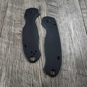 May include: Two black knife handle scales with multiple holes and a textured surface. The scales are made of a durable material and are designed to fit a folding knife. The scales are shown on a wooden surface.