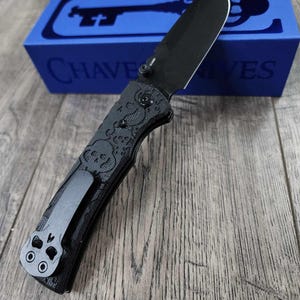 May include: A black folding knife with a skull pattern on the handle and a black pocket clip. The knife is in its closed position and is sitting on a wooden surface. The knife is in a blue box with the word "CHAVE KNIVES" printed on it.