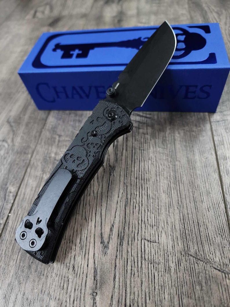 May include: A black folding knife with a skull pattern on the handle and a black pocket clip. The knife is in its closed position and is sitting on a wooden surface. The knife is in a blue box with the word "CHAVE KNIVES" printed on it.