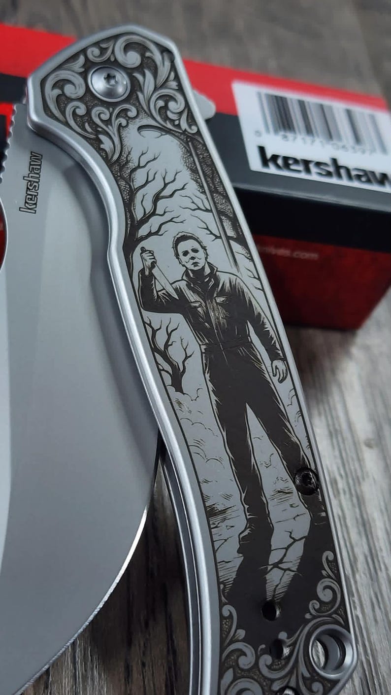 Pu&ograve; includere: Coltello pieghevole argentato con un'incisione dettagliata di una figura che tiene un coltello, su uno sfondo di albero e fogliame. La lama del coltello &egrave; visibile e il marchio "Kershaw" &egrave; stampato sulla lama.