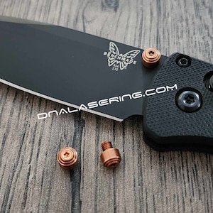 May include: Three copper-colored screws for a Benchmade 535 knife. The screws have a star-shaped head and are on a wooden surface.