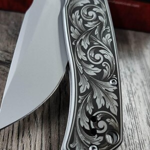 May include: A partially open folding knife with a silver blade and a handle with an engraved floral pattern. The handle has a dark background with a silver floral design. The knife's blade is visible, showing its sharp edge.