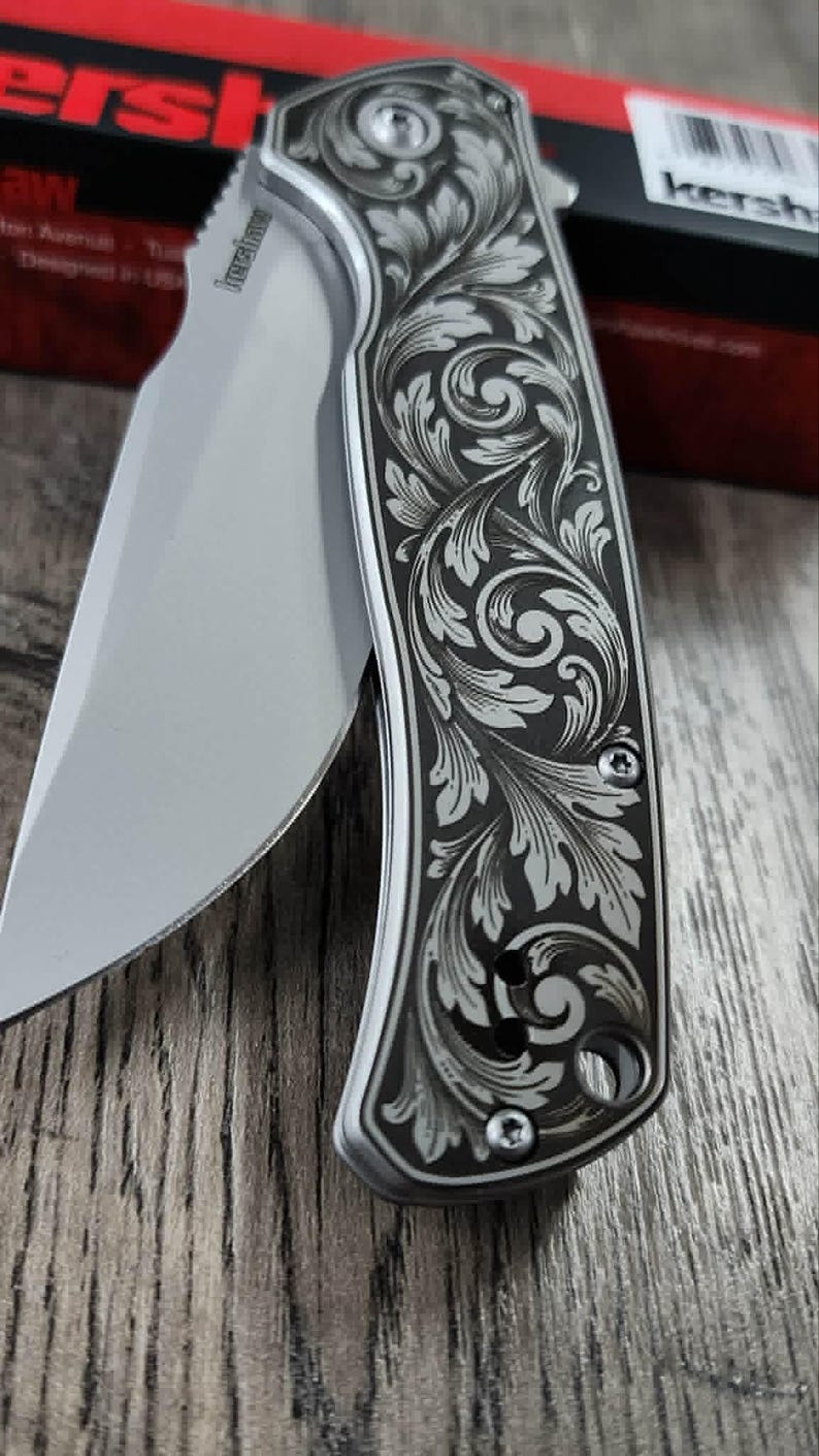 May include: A partially open folding knife with a silver blade and a handle with an engraved floral pattern. The handle has a dark background with a silver floral design. The knife's blade is visible, showing its sharp edge.