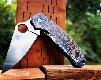 May include: A folding pocket knife with a silver blade featuring a circular thumb hole and a Spyderco logo. The handle has a dark blue and copper-toned topographic map pattern with the text "LASERLAB.COM" engraved along the side of the grip.