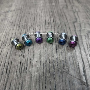 May include: A collection of six metal plugs with colorful, iridescent ends. The plugs are arranged in a row and feature a variety of colors, including green, blue, purple, and pink. The plugs are on a gray wooden surface.