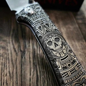 May include: A silver pocket knife with a detailed engraved design featuring a skull and intricate patterns. The handle is engraved with the text "DNALASERING.COM".