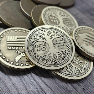 以下が含まれることがあります： 精巧なデザインのブロンズ色の金属コイン。1つのコインには生命の樹と「RULE #9 NEVER GO ANYWHERE WITHOUT A KNIFE」の文字が描かれています。他のコインにはアメリカ国旗と「MADE IN」の文字が表示されています。