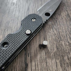 Puede incluir: Un cuchillo plegable negro con una hoja dentada y un mango negro texturizado. Dos pequeños tornillos plateados están junto al cuchillo sobre una superficie de madera.