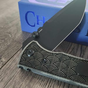 May include: Close-up of a black folding knife with a patterned handle and a partially open blade. The blade is marked with "ELMAX". The knife is set against a blue box with the text "Chaos Knives".