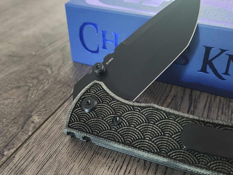 May include: Close-up of a black folding knife with a patterned handle and a partially open blade. The blade is marked with "ELMAX". The knife is set against a blue box with the text "Chaos Knives".