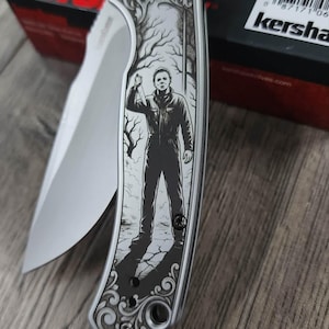 Pu&ograve; includere: Un coltello pieghevole argentato con una lama e un manico con un'immagine incisa di una figura che tiene un coltello. Il manico ha un design decorativo. Il marchio "Kershaw" &egrave; visibile.
