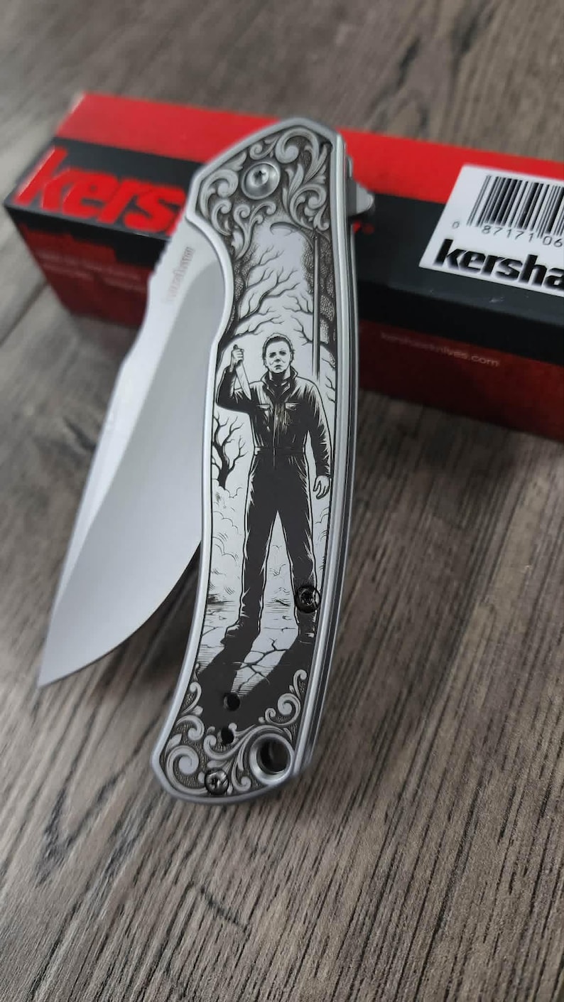 Pu&ograve; includere: Un coltello pieghevole argentato con una lama e un manico con un'immagine incisa di una figura che tiene un coltello. Il manico ha un design decorativo. Il marchio "Kershaw" &egrave; visibile.
