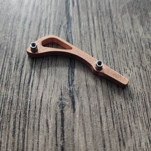 May include: A copper-colored metal component with two small silver-colored screws. The piece has a curved, open design with a rounded end and a narrower, pointed end. The item is set against a wooden surface.
