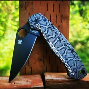 Puede incluir: Un cuchillo de bolsillo negro con un patrón de piel de serpiente gris y blanco en el mango. El cuchillo tiene un logotipo de Spyderco en la hoja y el texto "Spyderco" y "www.lasergrips.com" en el mango.