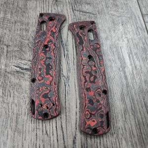 Puede incluir: Dos cachas de cuchillo con patrón negro y rojo. Las cachas tienen un patrón de remolino de negro y rojo.