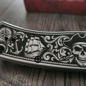 Puede incluir: Una navaja plateada con un mango negro con un dise&ntilde;o de tem&aacute;tica pirata. El dise&ntilde;o incluye una rosa de los vientos, un ancla, un velero y una calavera con un sombrero de pirata. El cuchillo tiene un borde decorativo.