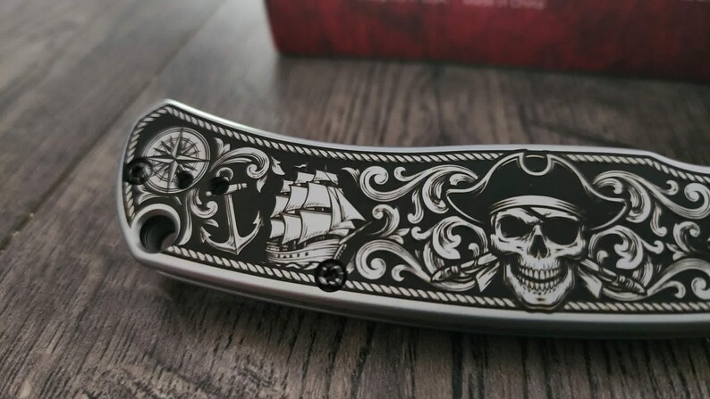 Puede incluir: Una navaja plateada con un mango negro con un dise&ntilde;o de tem&aacute;tica pirata. El dise&ntilde;o incluye una rosa de los vientos, un ancla, un velero y una calavera con un sombrero de pirata. El cuchillo tiene un borde decorativo.