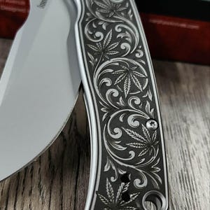 May include: A silver and black folding knife with a detailed engraved design of cannabis leaves and swirling patterns. The blade is partially open, revealing the sharp edge. The knife is set against a wooden surface.