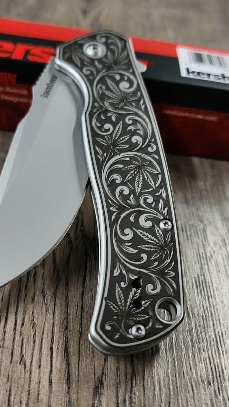 May include: A silver and black folding knife with a detailed engraved design of cannabis leaves and swirling patterns. The blade is partially open, revealing the sharp edge. The knife is set against a wooden surface.