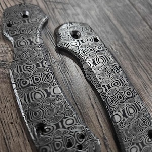 May include: Two knife handles with a distinctive swirling pattern in grey and black. The handles feature screw holes and a textured finish. The design is reminiscent of concentric circles and organic forms, producing a visually striking effect.