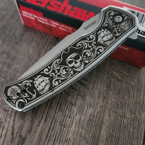 Puede incluir: Cuchillo plegable plateado con dise&ntilde;o de tem&aacute;tica pirata. La hoja presenta un cr&aacute;neo grabado con sombrero de pirata, un barco, un ancla y una rosa de los vientos. El cuchillo est&aacute; sobre una caja roja y negra.