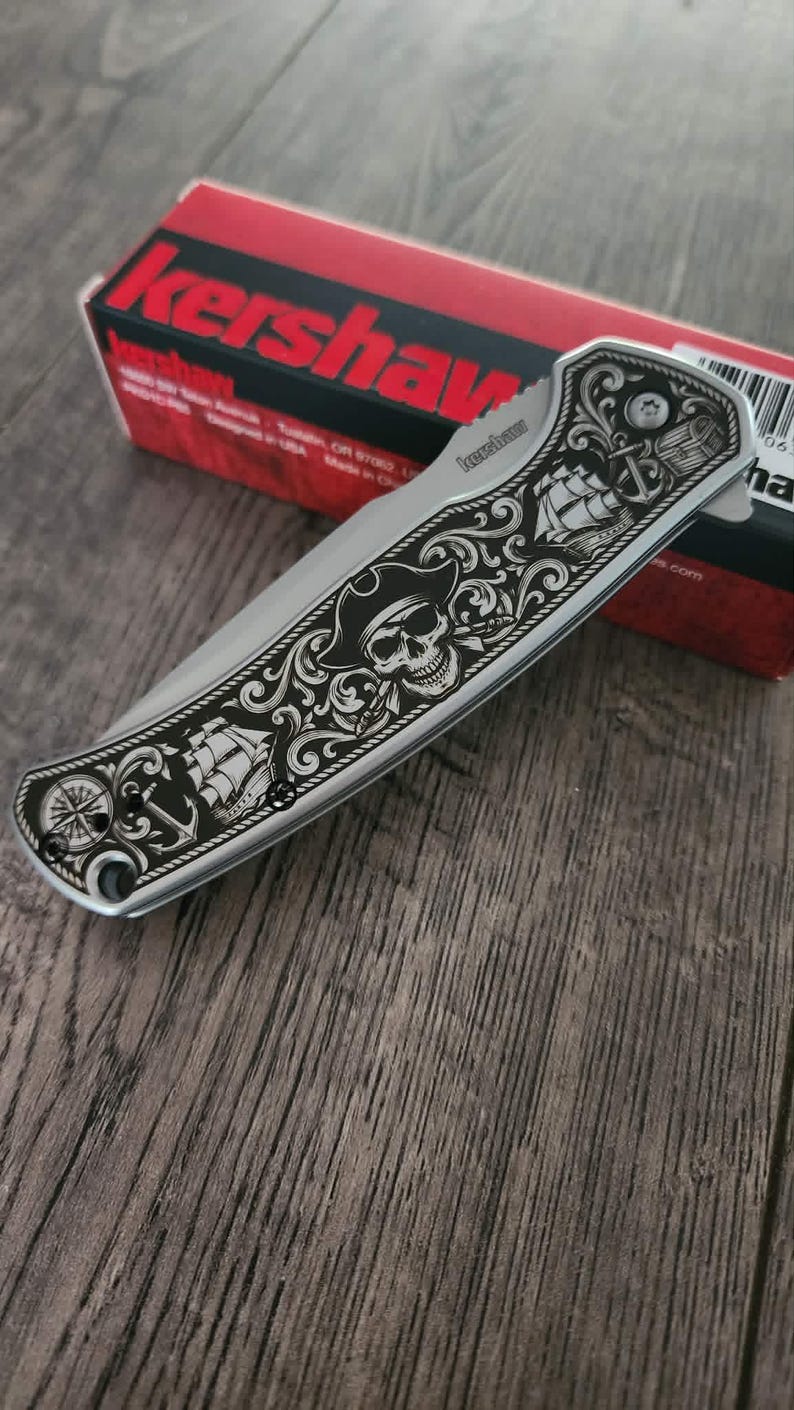 Puede incluir: Cuchillo plegable plateado con dise&ntilde;o de tem&aacute;tica pirata. La hoja presenta un cr&aacute;neo grabado con sombrero de pirata, un barco, un ancla y una rosa de los vientos. El cuchillo est&aacute; sobre una caja roja y negra.