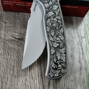 May include: A silver folding knife with an open blade. The handle has an engraved floral pattern. The knife is on a wooden surface, with a red and black box behind it. The brand name "Kershaw" is visible on the knife and box.