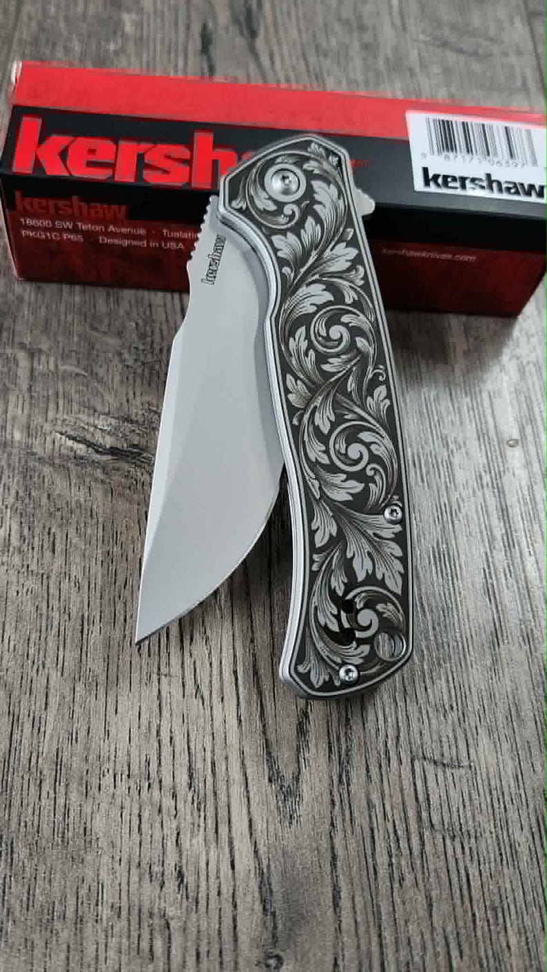May include: A silver folding knife with an open blade. The handle has an engraved floral pattern. The knife is on a wooden surface, with a red and black box behind it. The brand name "Kershaw" is visible on the knife and box.