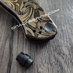 Puede incluir: Primer plano de una navaja de bolsillo de color dorado con un intrincado patrón floral grabado en el mango. La navaja tiene un mango negro y un botón negro en el lateral. También se puede ver una pequeña pieza de plástico negro en la imagen.