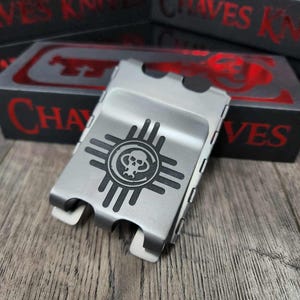 Peut inclure: Un porte-billets en métal argenté avec un motif graphique noir représentant un crâne dans un symbole solaire. Le clip a deux languettes blanches et est posé sur une surface en bois. Le texte "CHAVES KNIVES" est visible en rouge sur une boîte noire en arrière-plan.