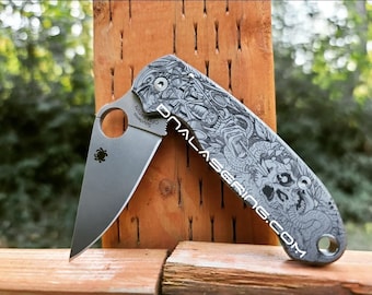 May include: A gray pocket knife with a black and white skull and snake design on the handle. The knife is partially open and the blade is visible. The knife is sitting on a wooden surface.