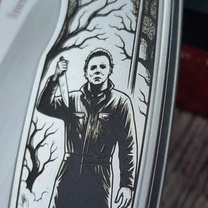 Pu&ograve; includere: Un coltello tascabile argentato con un'incisione dettagliata di una figura che tiene un coltello, sullo sfondo di alberi spogli. La lama del coltello &egrave; visibile e l'impugnatura presenta il marchio Kershaw.