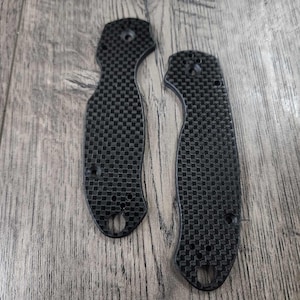 Puede incluir: Dos cachas de cuchillo de fibra de carbono negra con un patrón de cuadros. Las cachas tienen una forma curva con agujeros para tornillos y un agujero para cordón. Diseñado para una navaja plegable.