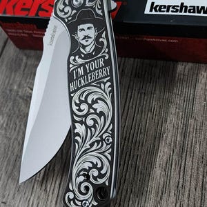 May include: A black and silver folding knife with a decorative handle featuring a portrait and the text "I'M YOUR HUCKLEBERRY". The blade is silver, and the knife is partially open. The knife is on a wooden surface.