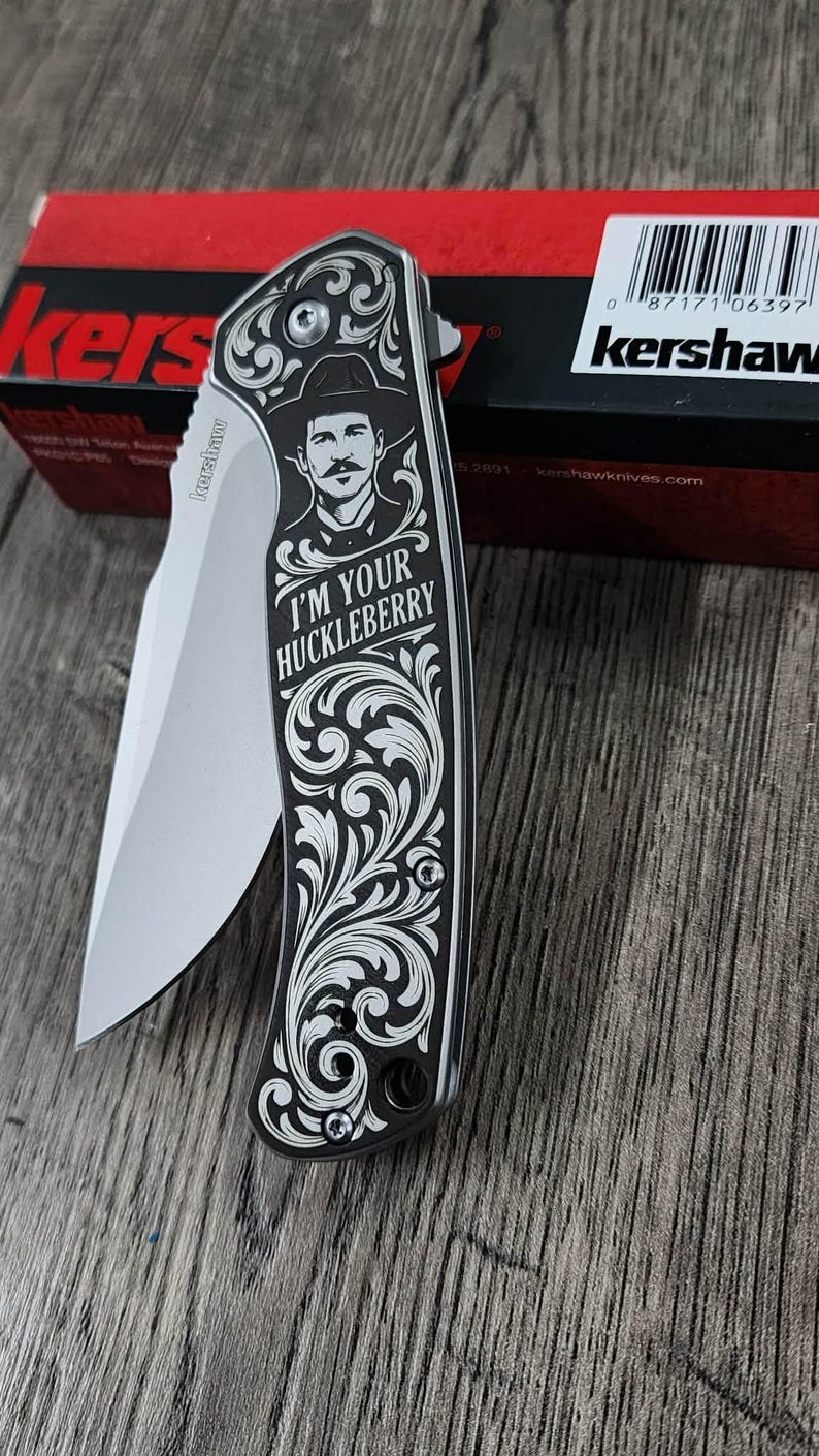 May include: A black and silver folding knife with a decorative handle featuring a portrait and the text "I'M YOUR HUCKLEBERRY". The blade is silver, and the knife is partially open. The knife is on a wooden surface.